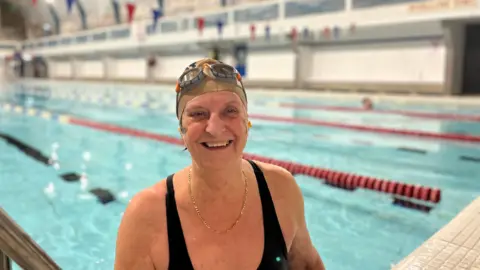 BBC A swimmer stands in the pool, smiling at the camera. She wears a swimming cap, googles, black swimsuit and gold necklace.
